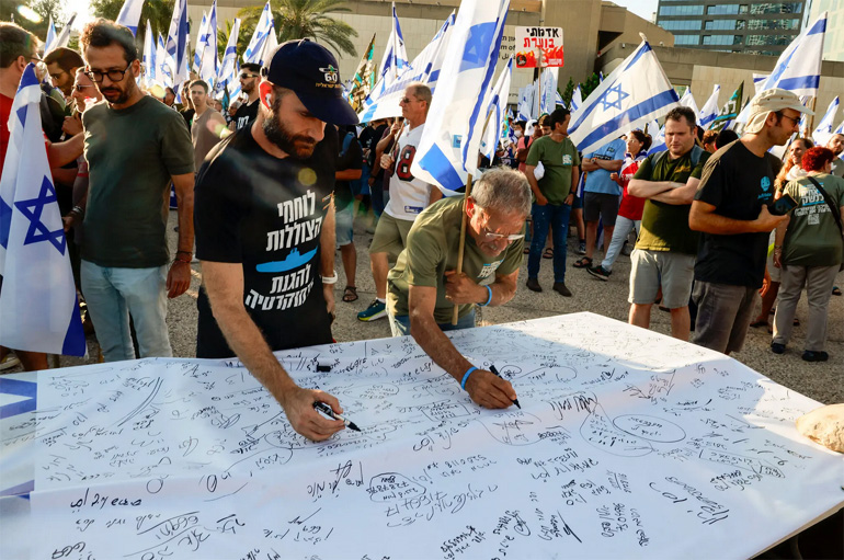  Ισραηλινοί έφεδροι στρατιωτικοί υπογράφουν μια μεγάλη διακήρυξη στο Τελ Αβίβ που ανακοινώνουν την αναστολή των εθελοντικών τους καθηκόντων, με άλλους διαδηλωτές στο βάθος να κρατούν ισραηλινές σημαίες